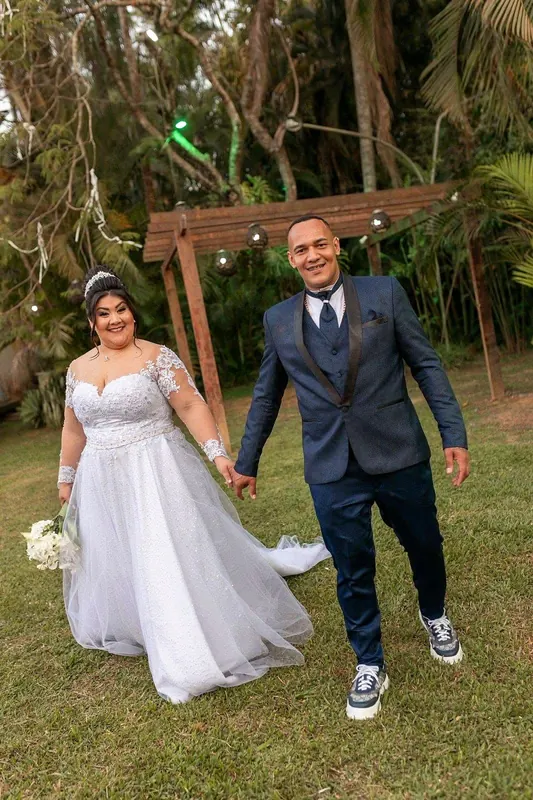 Noivos caminhando de mãos dadas no gramado, com o pergolado e o bosque ao fundo